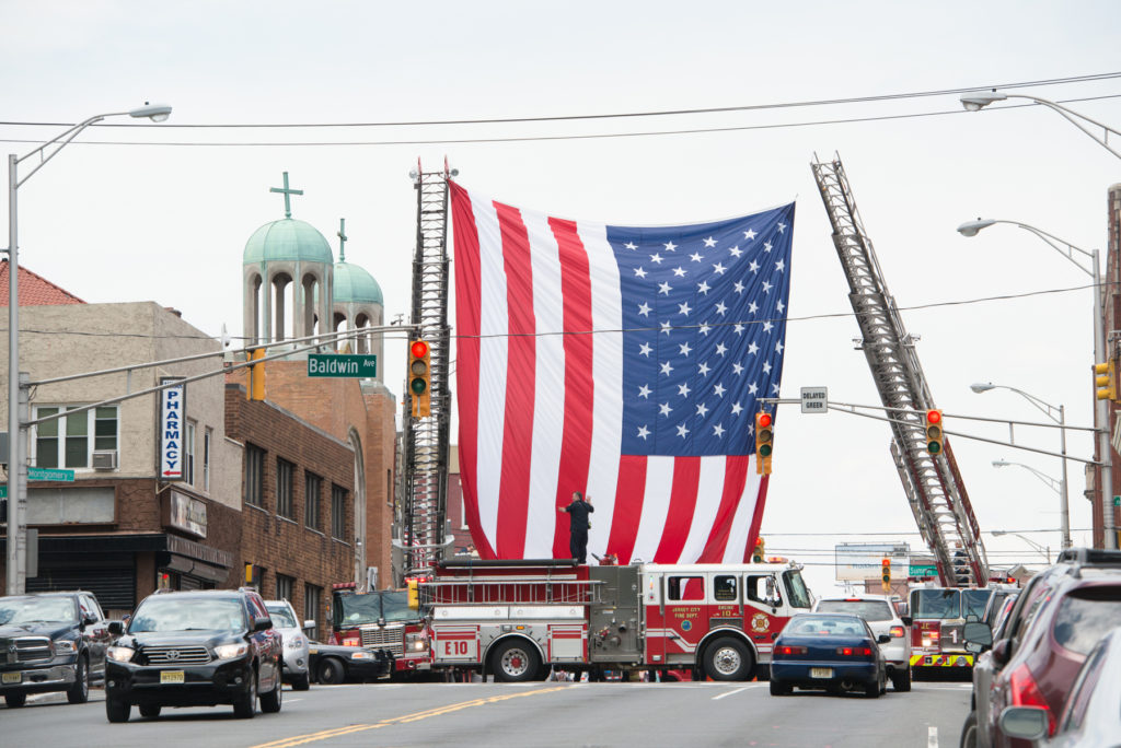The Jersey City Fire Department prepares to welcome home 160 soldiers of the New Jersey Army National Guard's Alpha Company, 2-113th Infantry Battalion, after a nine-month deployment in the Persian Gulf, by displaying a giant American flag between two fire ladder trucks in the middle of Montgomery Street on Thursday, June 25, 2015. Four buses transported the citizen soldiers to the National Guard Armory for a ceremony attended by their families. Reena Rose Sibayan | The Jersey Journal